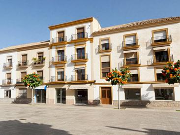Calle soleada con edificios de cinco plantas, bajos comerciales y naranjos, imagen de ambiente creada con IA
